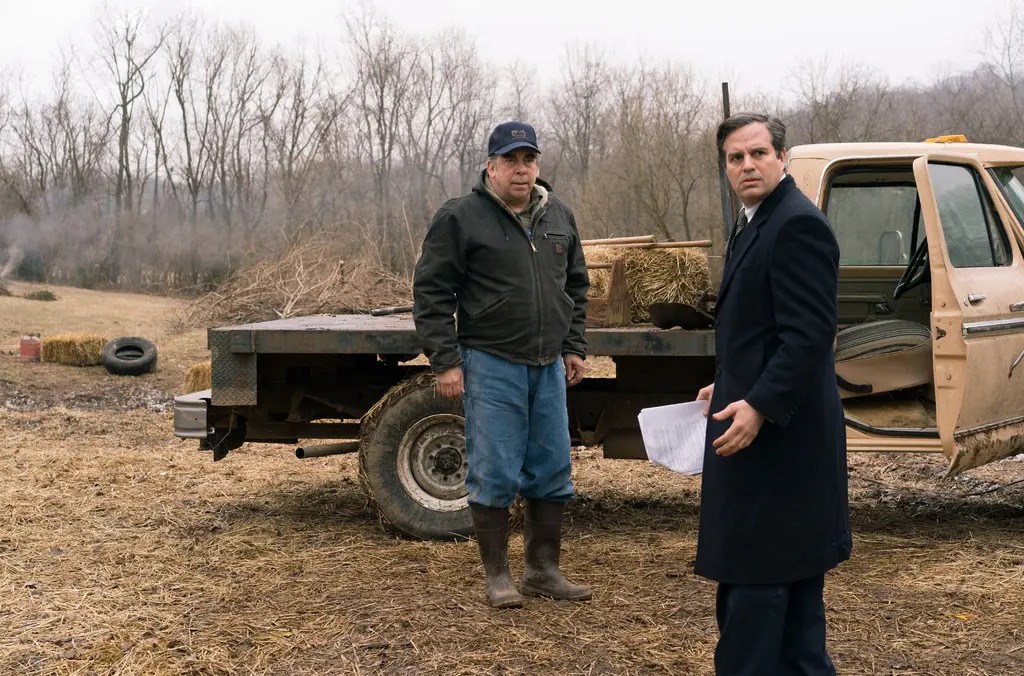 A man in a dark overcoat looking over his shoulder with a look of concern next to a man standing beside a flatbed truck in Dark Waters.