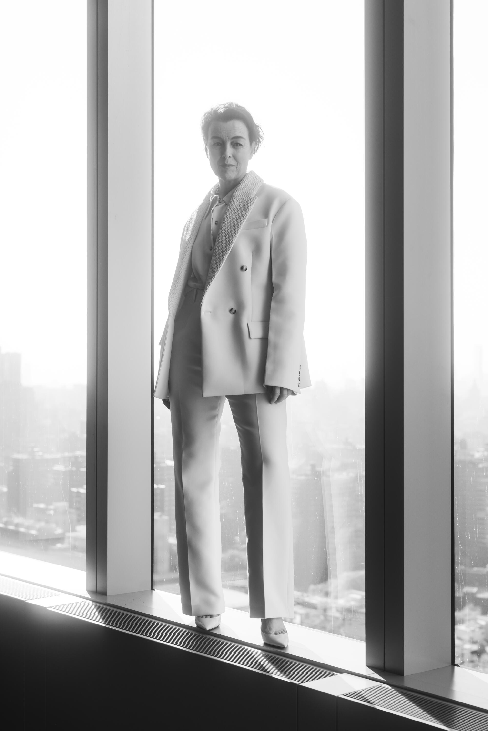 A black and white photo of a woman in a white suit standing on the ledge of a glass window.