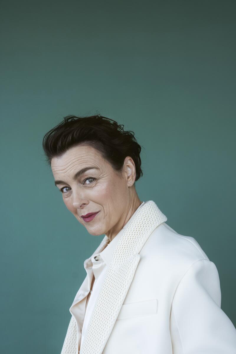 A woman with short hair in a white suit turns her head to smile at the camera.