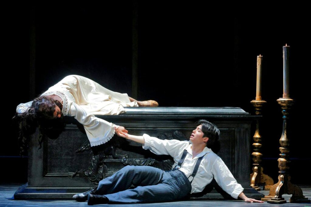 A young woman lies on a carved casket, reaching down to the young man on the floor next to it