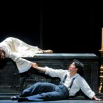 A young woman lies on a carved casket, reaching down to the young man on the floor next to it