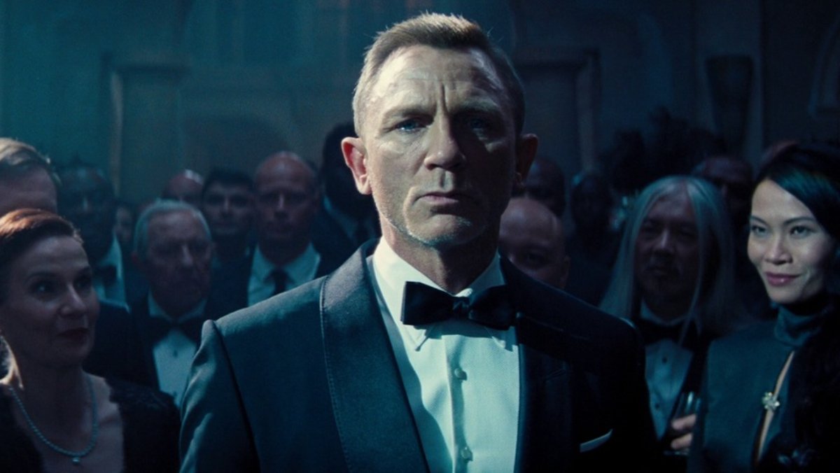 Daniel Craig wearing a suit with a bowtie staring ahead with many people looking at him from behind.