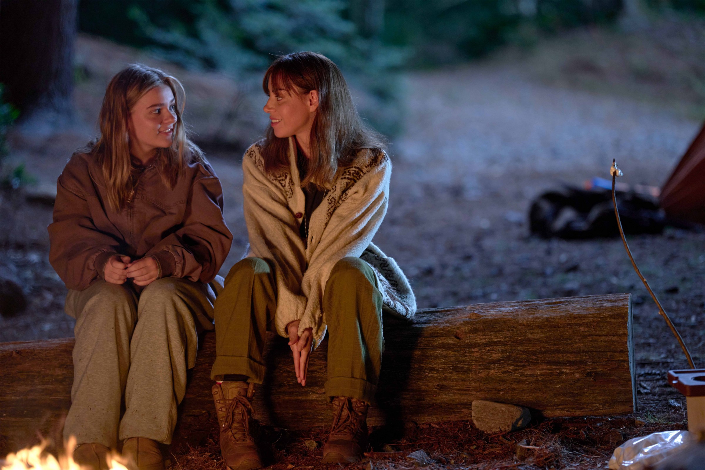 Elliott, a blonde teenage girl played by Maisy Stella, sits next to her older self, played by Aubrey Plaza, on a log by a campfire in My Old Ass