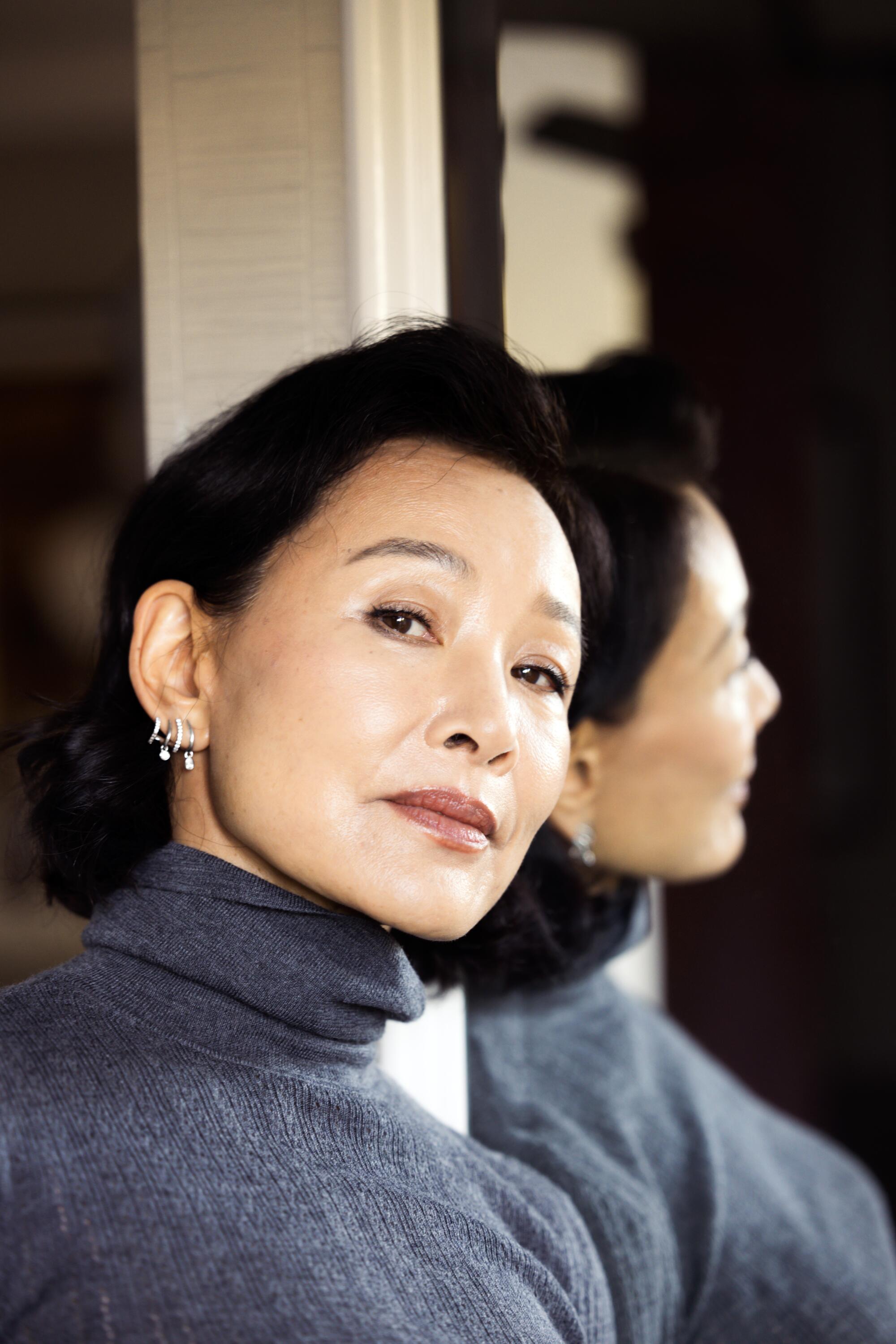 A woman in a grey turtle neck leans against a mirror