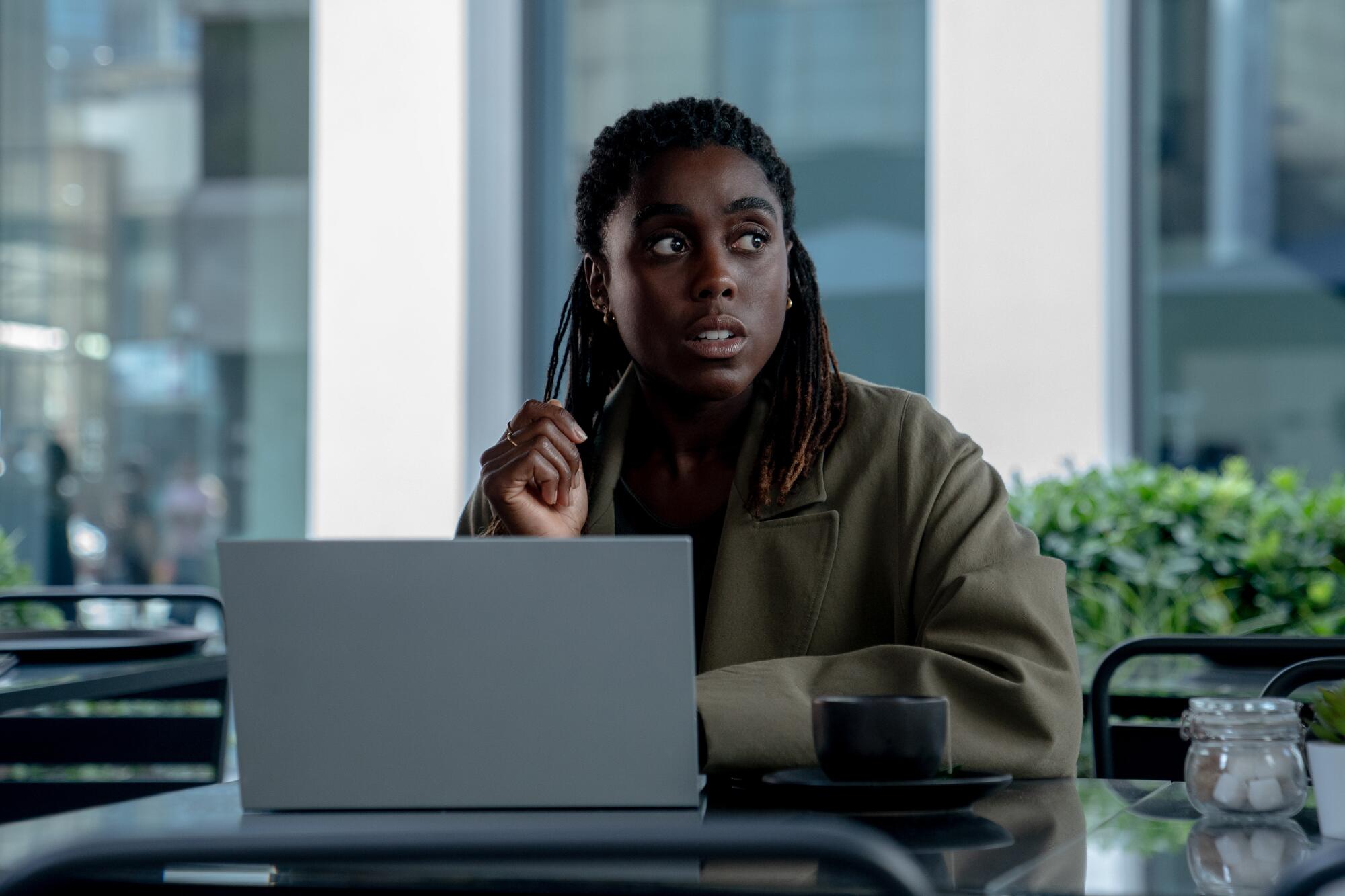 Lashana Lynch in a green jacket sitting in front of a silver laptop looks to the side.