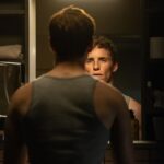 Eddie Redmayne in a gray tank top standing at a mirror looking at his reflection.