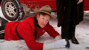 Conan O'Brien, dressed as a Canadian Mountie, lies in the snow caressing a woman's black boot.