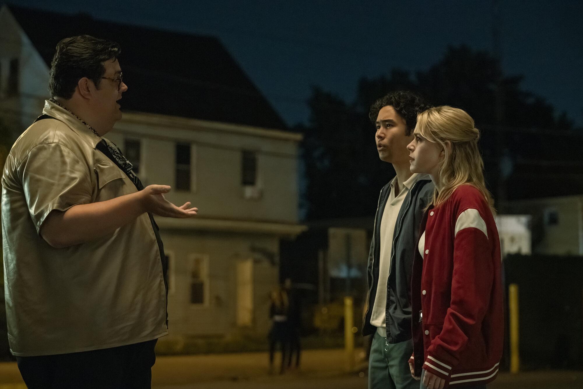 A guy in a tan shirt holds his hand out and looks at a couple speaking to him.