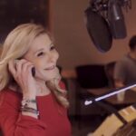 Pamela Hayden, a woman with blonde hair wearing a red shirt, in a recording studio holding an earphone to her ear