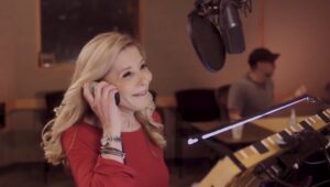 Pamela Hayden, a woman with blonde hair wearing a red shirt, in a recording studio holding an earphone to her ear