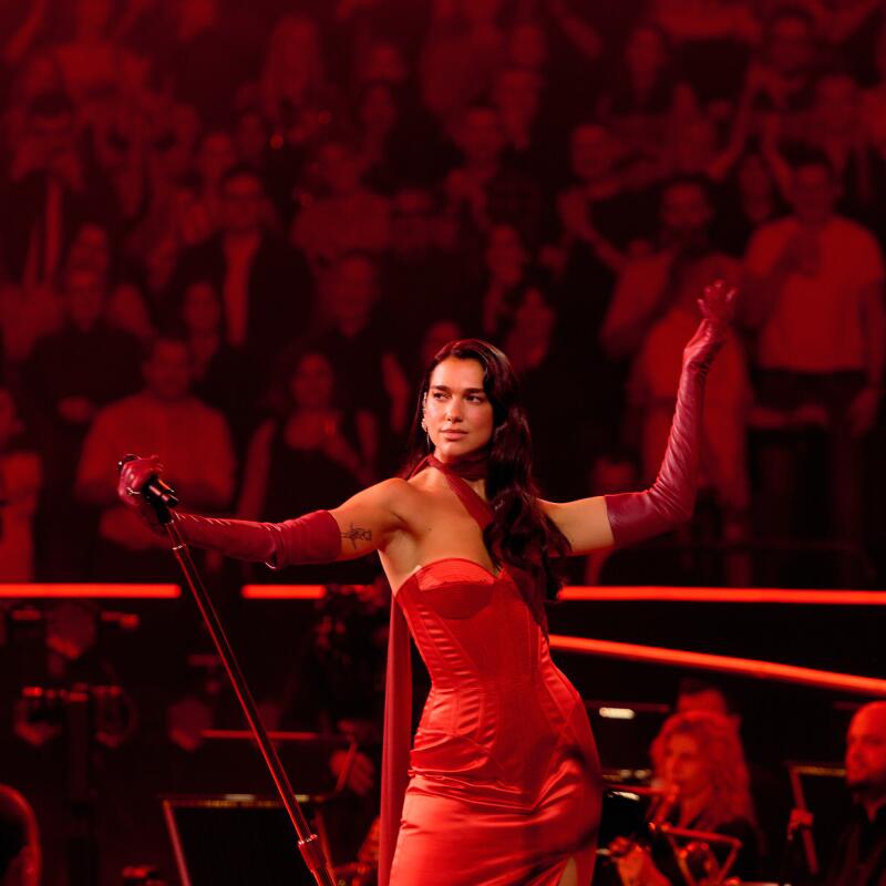 A woman in a red strapless gown holds a mic stand an lifts a red gloved hand in the air.