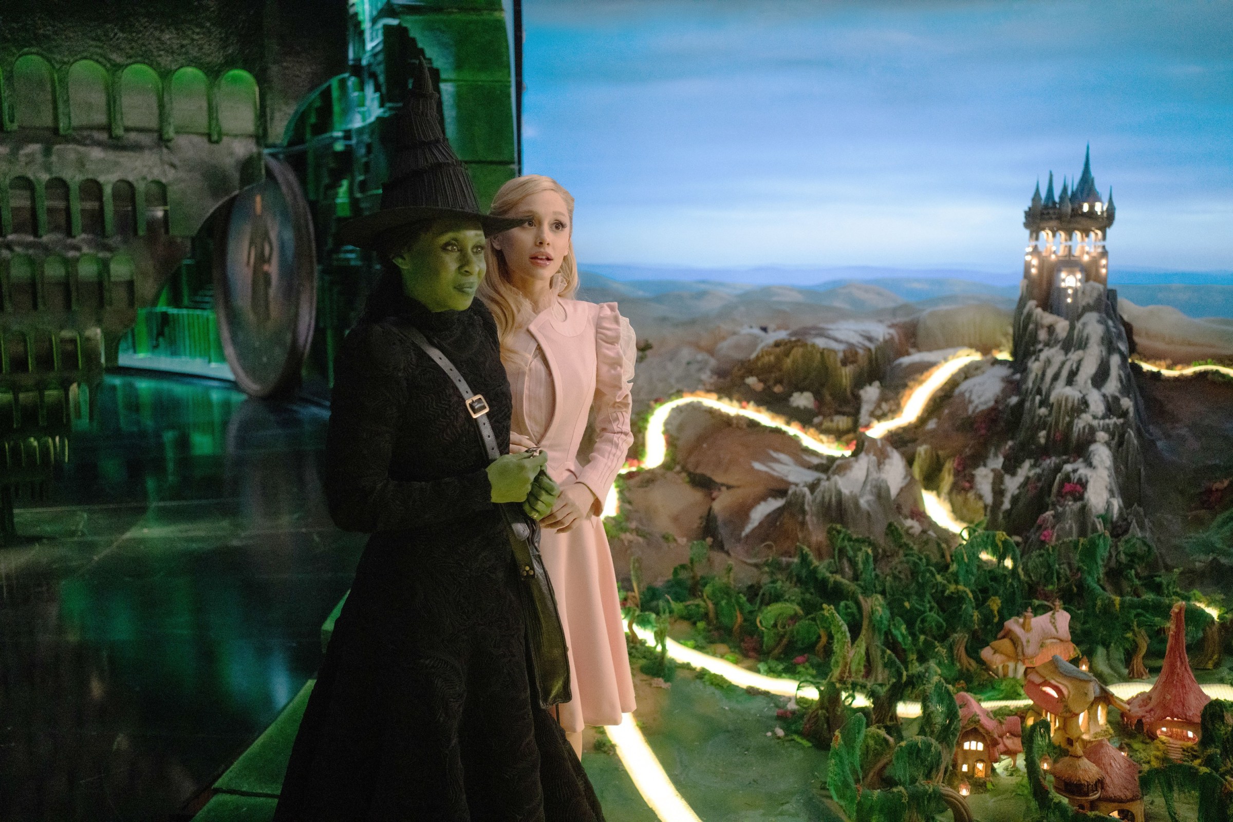 The green-skinned witch Elphaba in black (Cynthia Erivo) and the blonde witch Glinda in pink (Ariana Grande) stand together in front of a room filled with a huge, elaborate model of Oz in Wicked