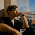 Cillian Murphy sits next to a window, his reflection in the glass, for a portrait.