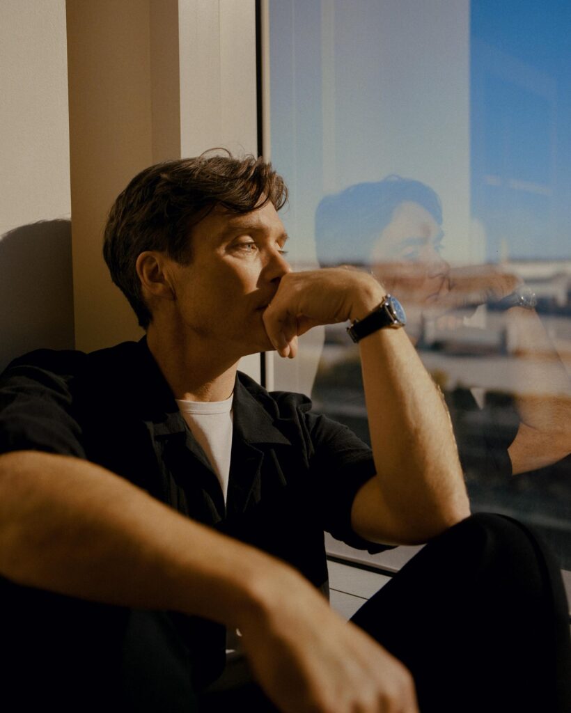 Cillian Murphy sits next to a window, his reflection in the glass, for a portrait.