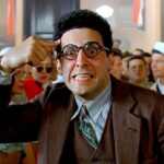 John Turturro wearing round glasses in a brown suit jacket pointing at his temple and gritting his teeth in Barton Fink.