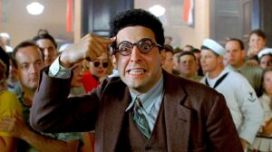 John Turturro wearing round glasses in a brown suit jacket pointing at his temple and gritting his teeth in Barton Fink.