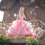 Glinda the Good (Ariana Grande, in a giant poofy pink ballgown, and carrying a wand with a sparkling silver-and-pink spangle on the tip) stands on a platform surrounded by metal lace balustrades and looks modest in Wicked