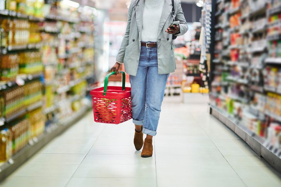 woman shopping in a grocery store with a basket in one hand and her phone in the other