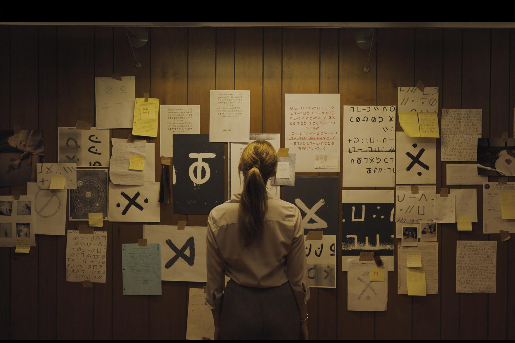 A woman stands facing a wall papers with symbols on them in "Longlegs."