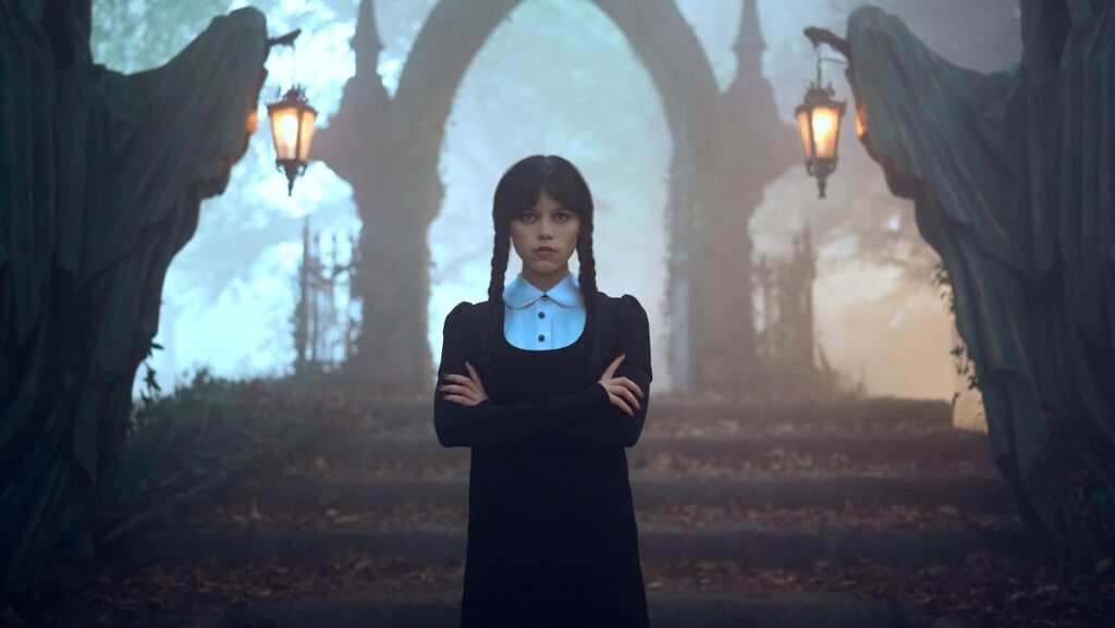 Jenna Ortega as Wednesday Addams with her arms crossed in front of stairs leading to an archway