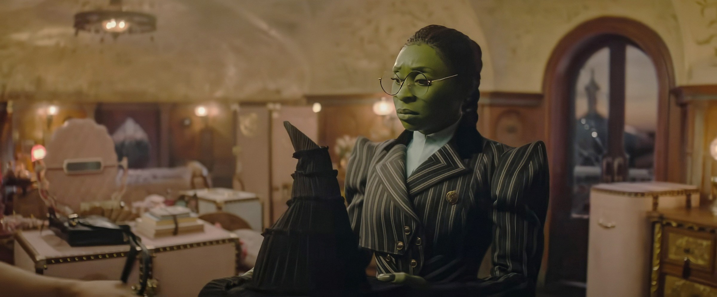 Elphaba (Cynthia Erivo), in a pinstriped suit with dramatic shoulder pads, forlornly regards the pointy black witch hat her supposed friend Glinda gave her in Wicked: Part I