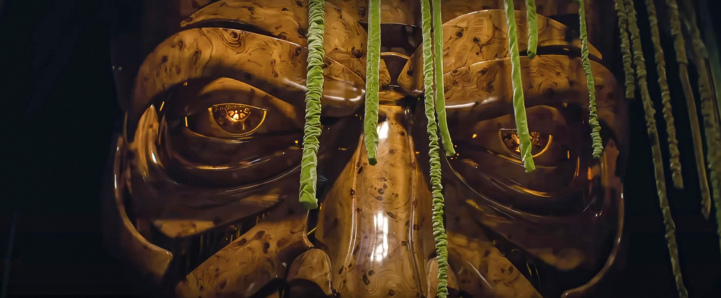 The giant talking mask of the Wizard of Oz, seemingly made of highly polished wood pieces assembled into an angry-looking face, seen in close-up in Wicked: Part I