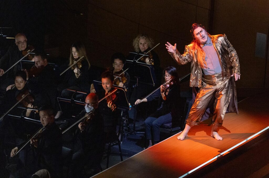 A costumed singer in a golden suit performs on a catwalk above string players of the L.A. Phil during "Das Rheingold."