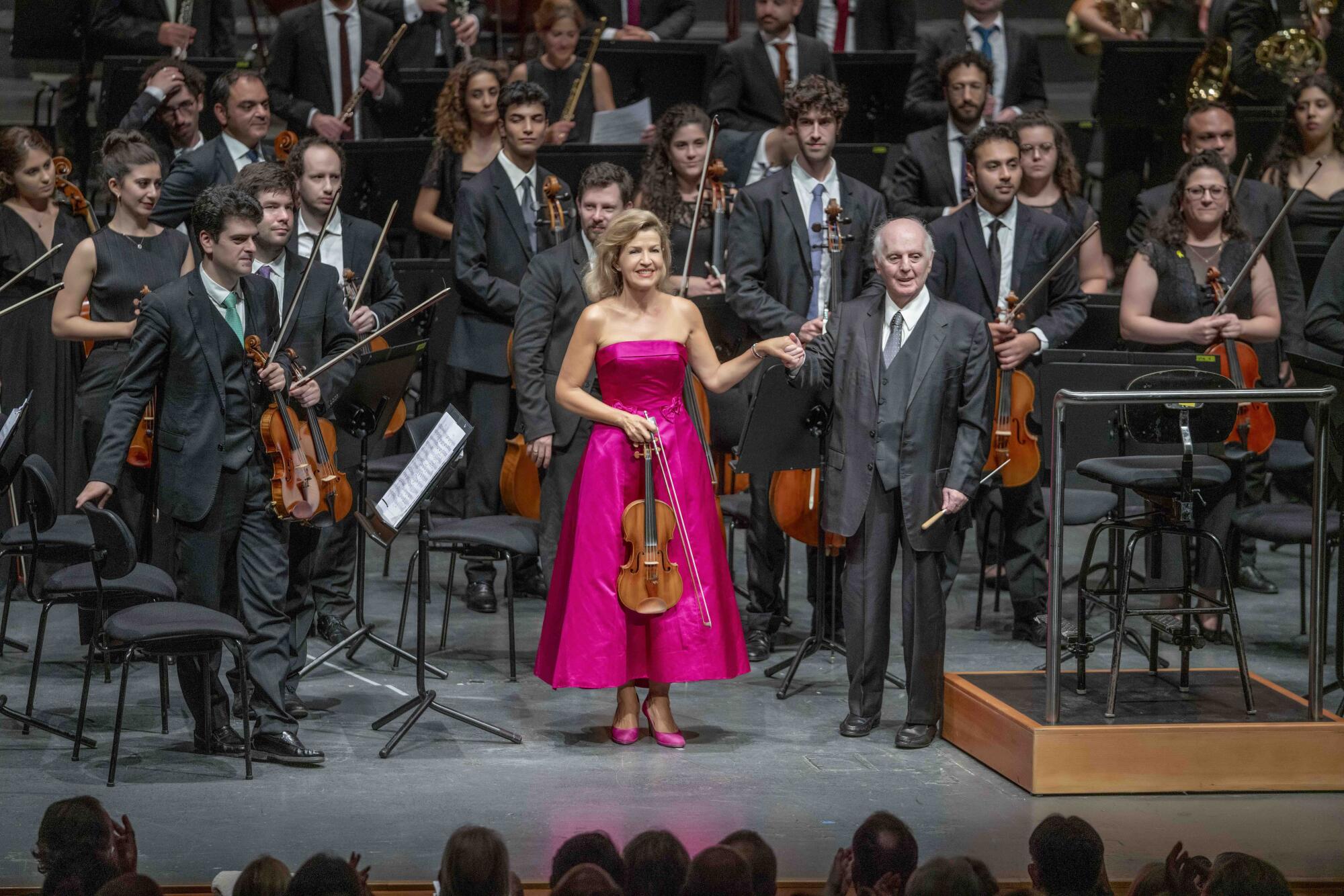 A conductor in a gray suit holds the hand of a violinist in a bright pink dress as young orchestra members stand behind them.