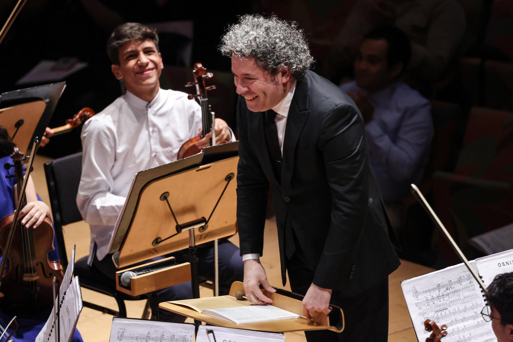 A conductor smiles and bows toward his orchestra as one of the young musicians grins.