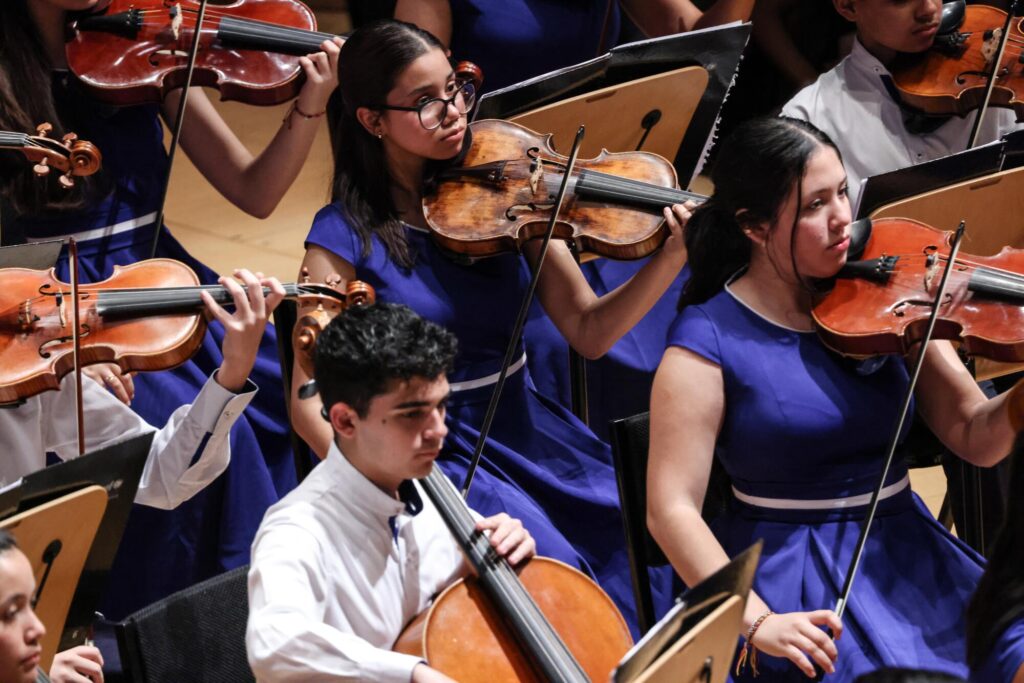 Two young women in purple dresses and a young man in a white shirt perform with the Children's Symphony of Venezuela.