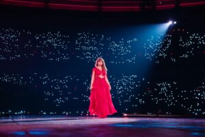 Taylor Swift performs in a floor length red dress in front of a black backdrop with white and red lights in it.