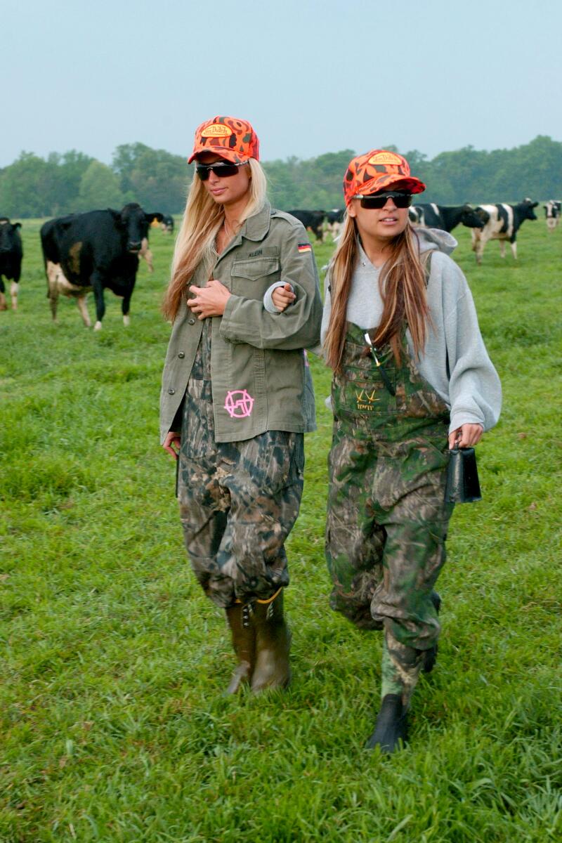 Two women in green camouflage overalls and orange hats walk through a field with cows in the background. 