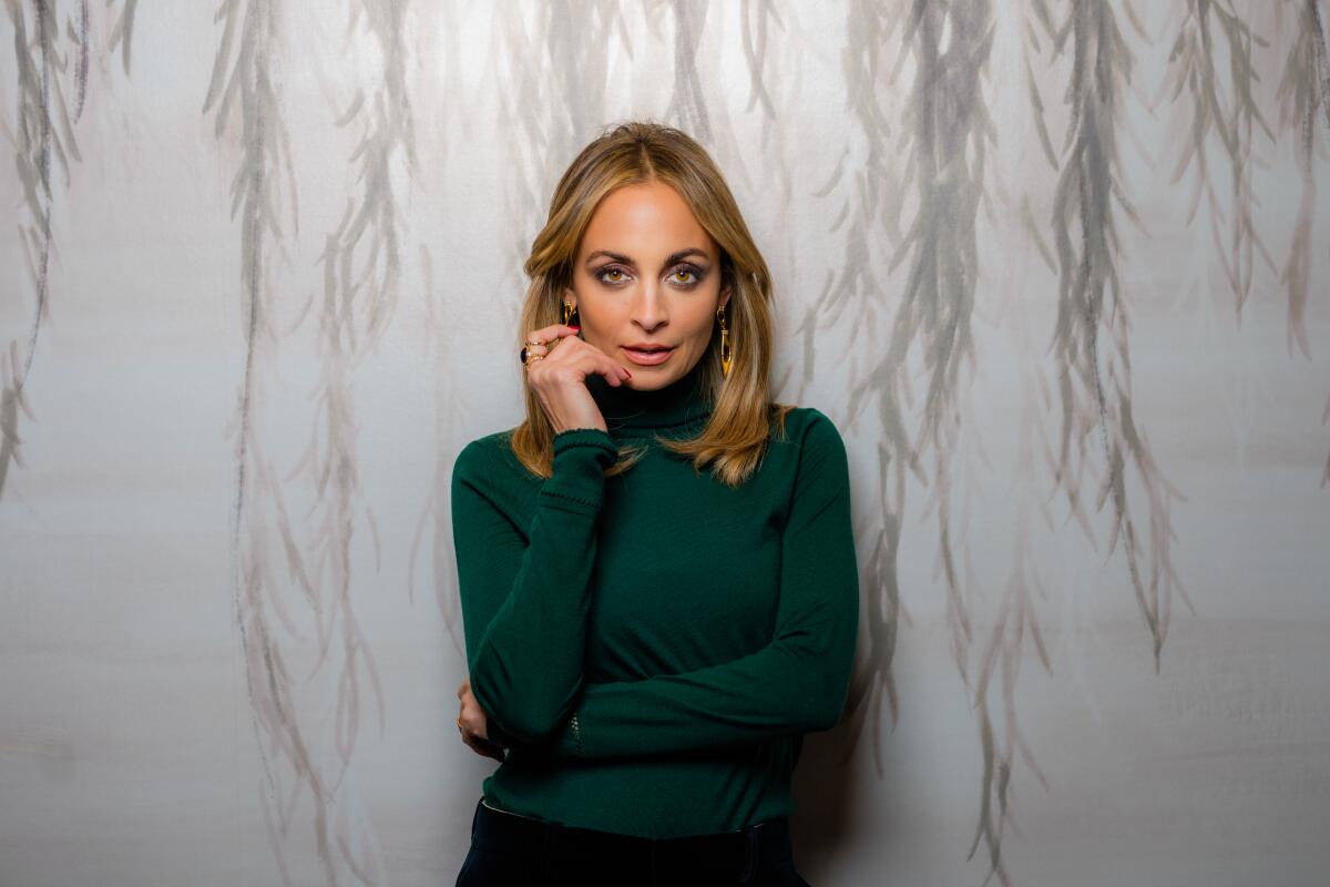 A woman with brown hair past her shoulders in a green turtleneck raises a hand near her chin.