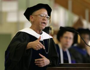 Nikki Giovanni, clad in academic robes and a hat, gestures with one hand as she recites a poem