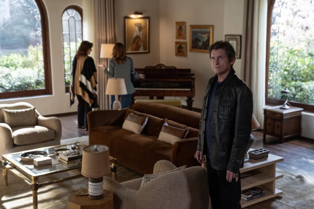 A man in a black leather jacket stands in a living room looking upward, with two women in the background