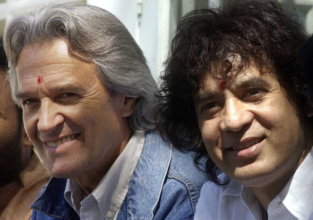 Guitarist John McLaughlin (L) and tabla maestro Ustad Zakir Hussain (R) listen to a journalist's question upon arrival at their hotel in Bangalore on Jan. 30, 2003. McLaughlin and Hussain will be performing in Bangalore 30 January with U.Srinivas and V.Silva Ganesh in a fusion of western jazz and traditional Indian styles. AFP PHOTO/INDRANIL MUKHERJEE (Photo by INDRANIL MUKHERJEE / AFP) (Photo by INDRANIL MUKHERJEE/AFP via Getty Images)