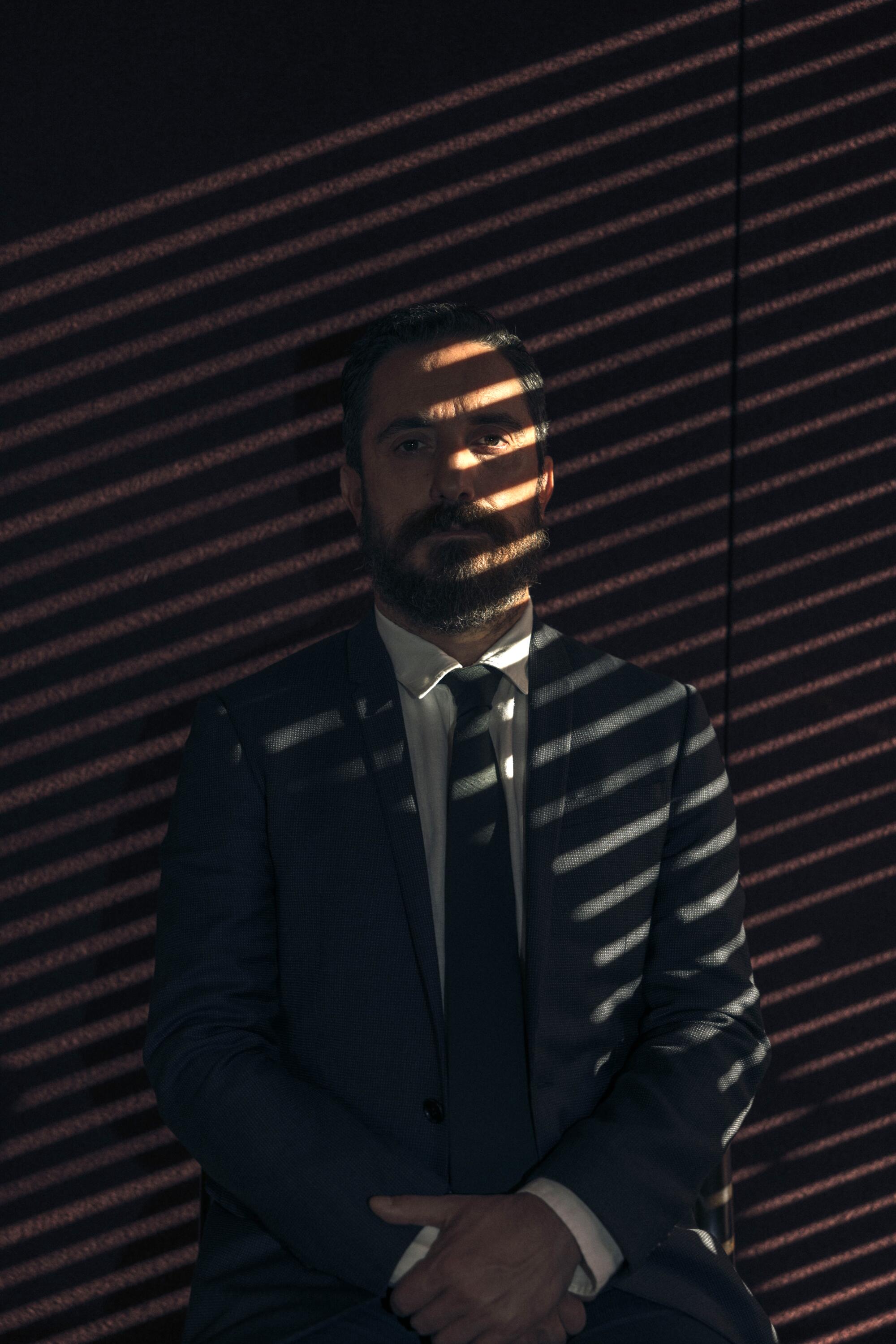 Director Pablo Larraín poses for a portrait near window blinds casting striped shadows across him.