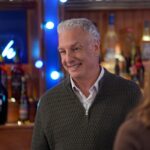 A man with white hair in a quarter-zip sweater standing near a bar with bottles of liquor.