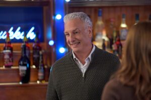 A man with white hair in a quarter-zip sweater standing near a bar with bottles of liquor.