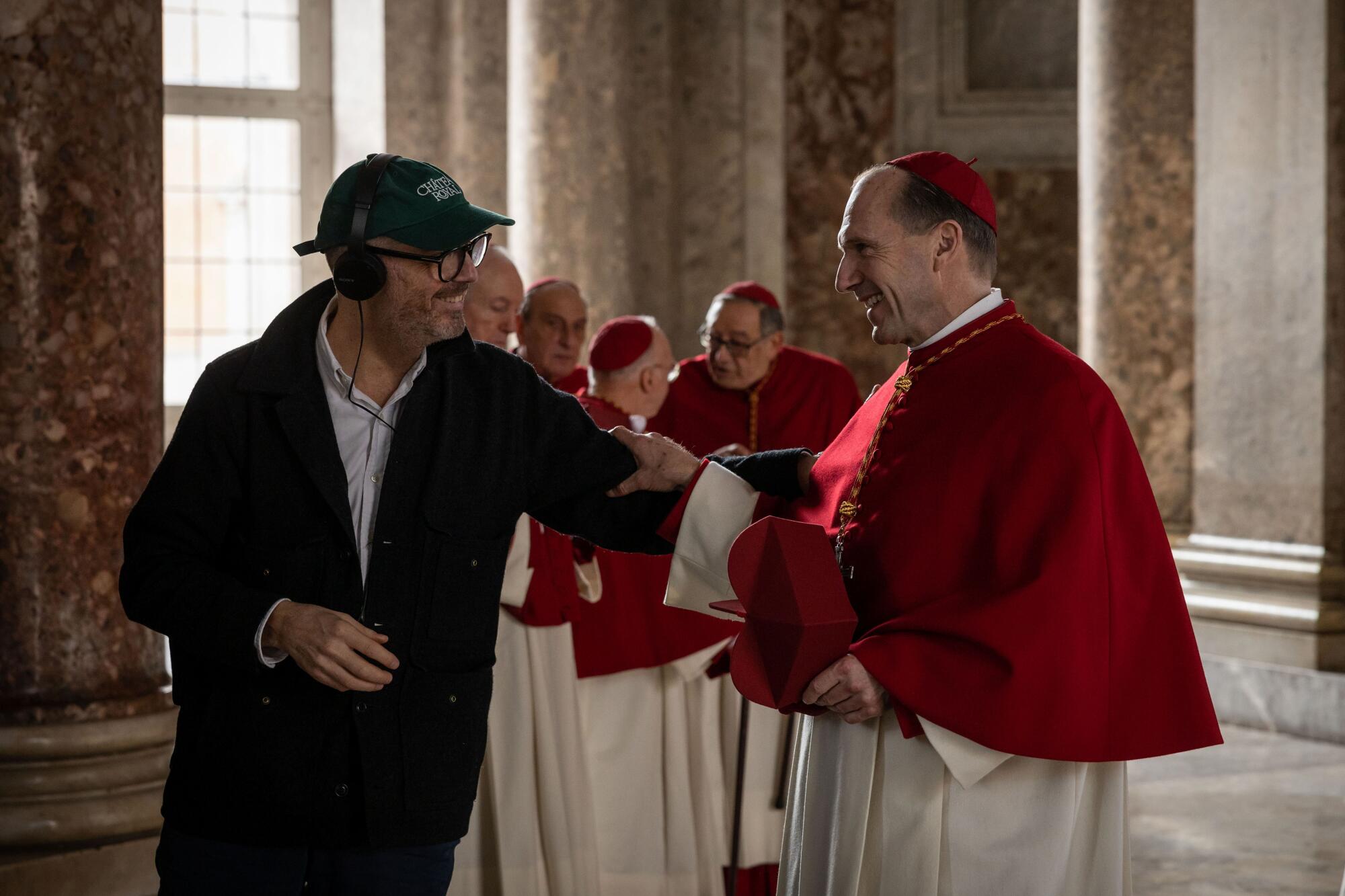 A man wearing a headset talks with a man in a cardinal's robes.