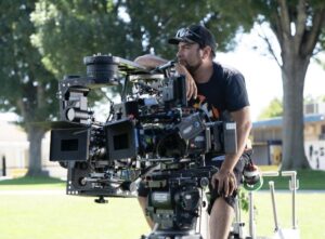 Diego Mariscal, in a black cap and T-shirt, sits on a camera dolly outdoors near tall trees.