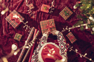 Top view of woman in red santa claus hat wrapping christmas gifts boxes for family sitting on fluffy plaid near christmas tree. Flat lay.