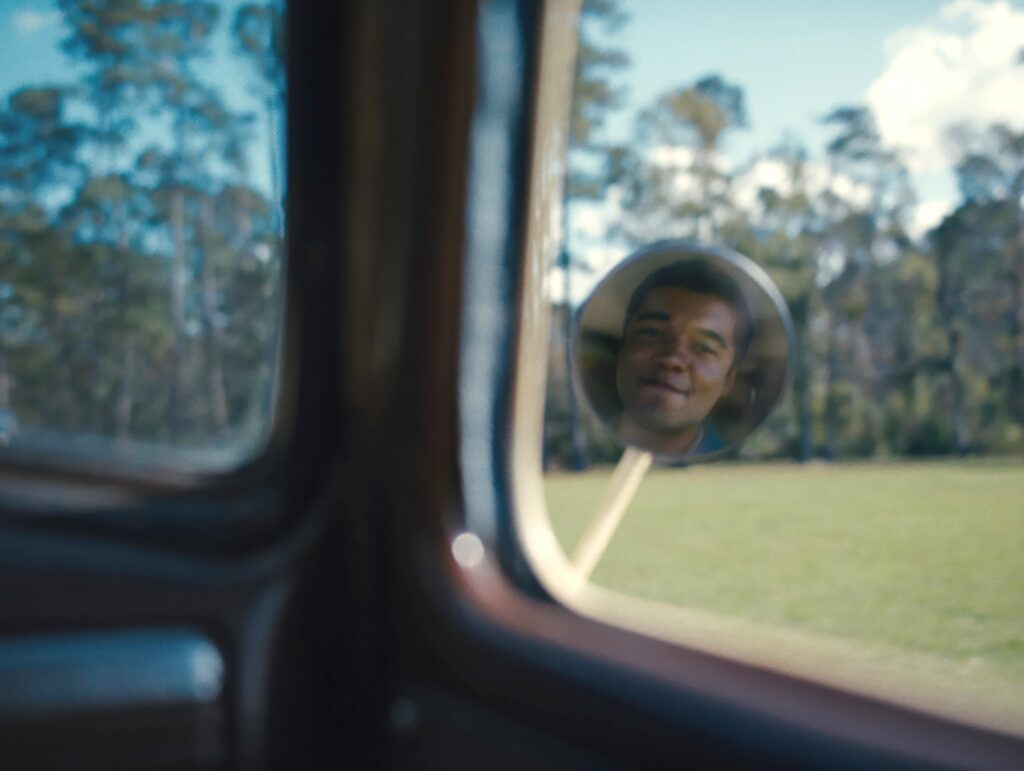 A young Black man’s smiling face is reflected in the wing mirror of an old car in Nickel Boys