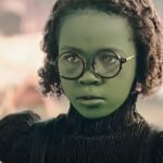 Wee li’l child Elphaba (Karis Musongole), a young green girl in black-rimmed glasses, glowers at off-screen bullies in Wicked: Part 1