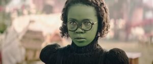 Wee li’l child Elphaba (Karis Musongole), a young green girl in black-rimmed glasses, glowers at off-screen bullies in Wicked: Part 1