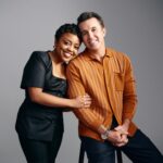 A woman in a black dress holds the arm of a man in an orange shirt sitting on a stool.