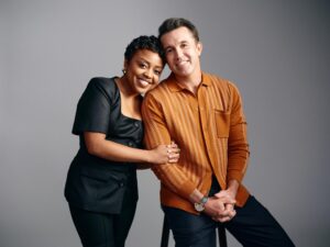 A woman in a black dress holds the arm of a man in an orange shirt sitting on a stool.