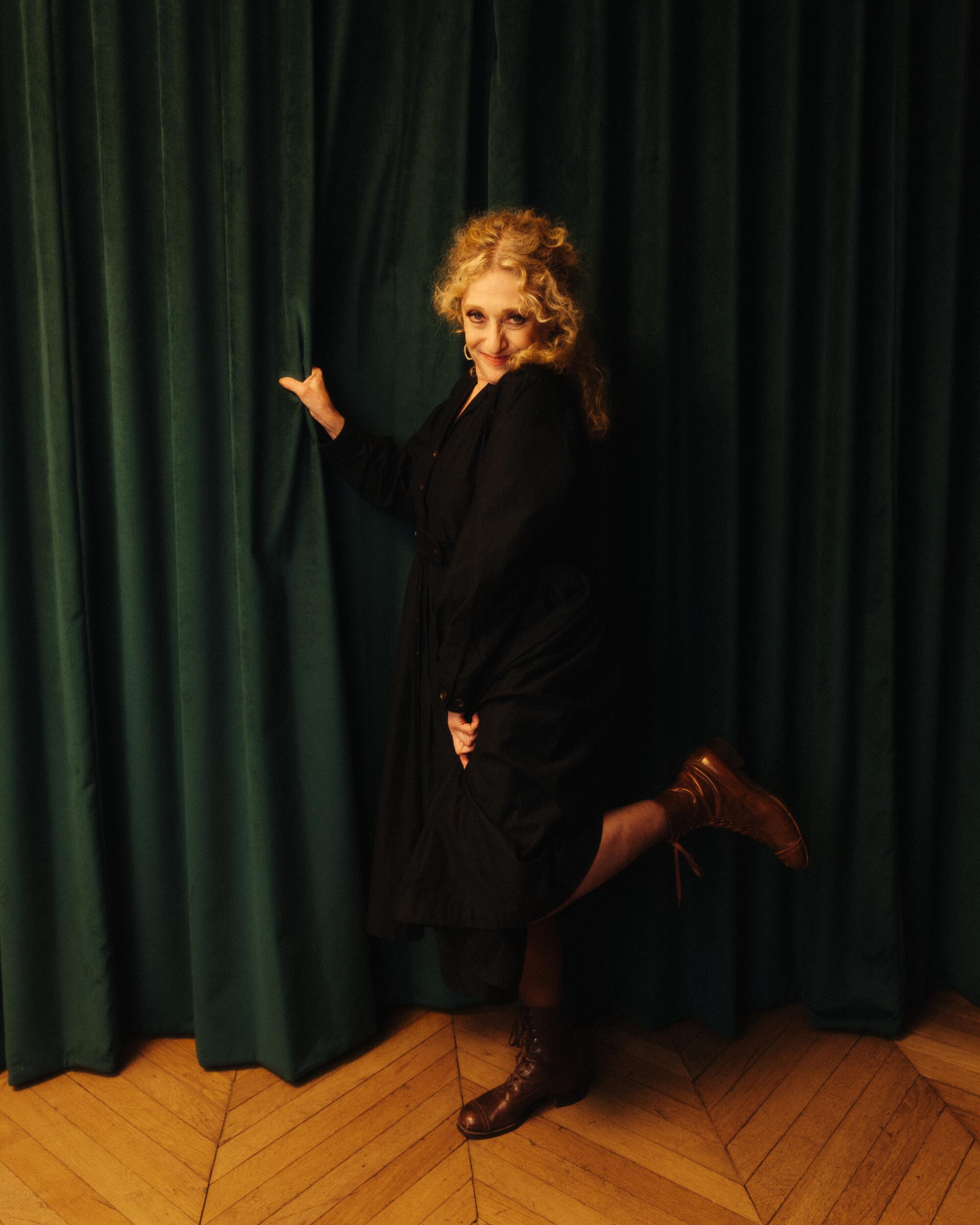 Carol Kane stands by long drapes and kicks up a heel in a portrait.