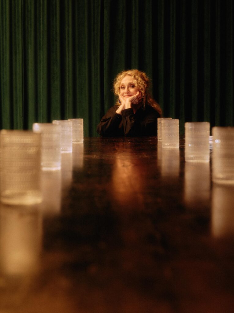 Carol Kane sits at the end of a long table, chin in her hands.