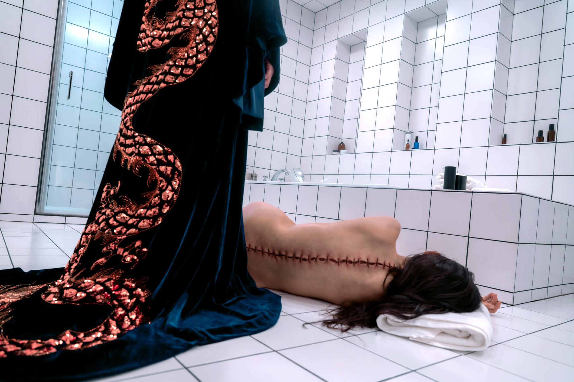 A figure in a robe stands over a woman lying on a bathroom floor with giant stitches down her spine in "The Substance."